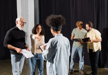 smiling multiracial woman pointing with hand near bearded art director and african american actress on theater stage,stock image